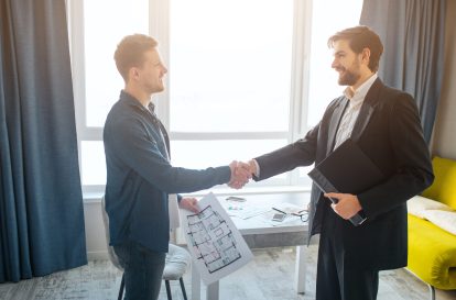 Young man and realtor buy or rent apartment. They shake hand having business deal. Positive and happy. Stand in one room. Daylight